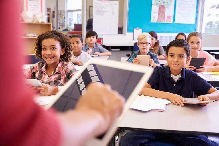 A photo of young elementary students looking at their teacher holding a netbook - image